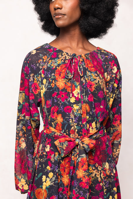 A woman wears the Ivy City Co Imogen Dress in Night Garden—a long sleeve, maternity-friendly floral maxi with vibrant red, orange, and yellow flowers. The dress features a waist bow and keyhole neckline with tie. Her face is partially out of frame.
