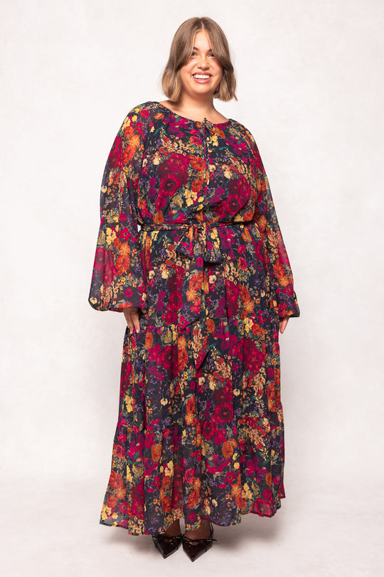 A smiling woman with short, light brown hair wears the Ivy City Co Imogen Dress in Night Garden—a maternity-friendly, long sleeve floral dress with a belted waist, featuring red, yellow, and orange flowers against a plain background.