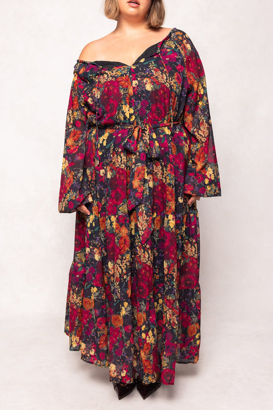 A woman wearing the Imogen Dress in Night Garden by Ivy City Co—a long sleeve, off-shoulder maxi dress adorned with vibrant red, yellow, and pink floral patterns on a dark background. This fall-ready floral dress features a belted waist and is designed to be effortlessly stylish and maternity friendly.