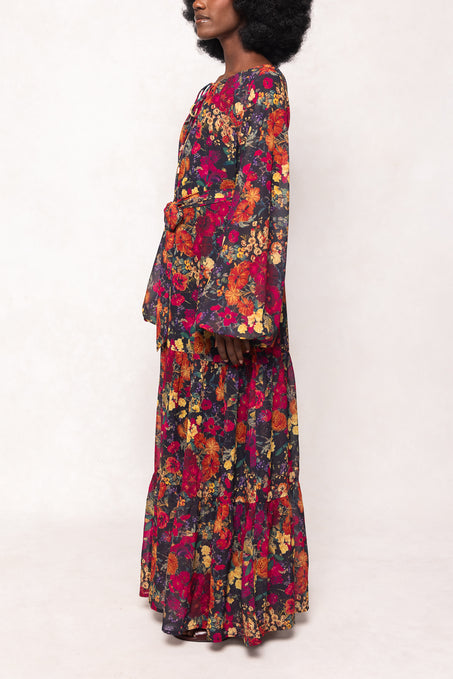 A woman stands in profile wearing Ivy City Co’s Imogen Dress in Night Garden, a long sleeve fall floral maxi with vibrant red, yellow, and orange patterns. Flared sleeves and a loose fit make it maternity friendly and comfortable.