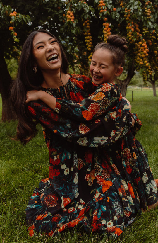 An adult and child wearing matching Ivy City Co Moody Floral dresses—including the Mini Imogen Dress—sit on orchard grass, laughing and hugging amid green trees with bright orange fruit.
