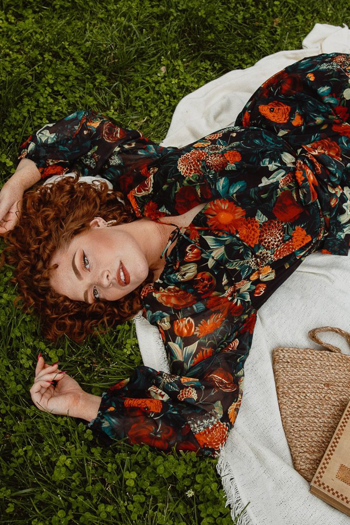 A woman with curly red hair lounges on green grass, smiling softly in the maternity-friendly Imogen Dress in Moody Floral by Ivy City Co. A woven bag is placed nearby.