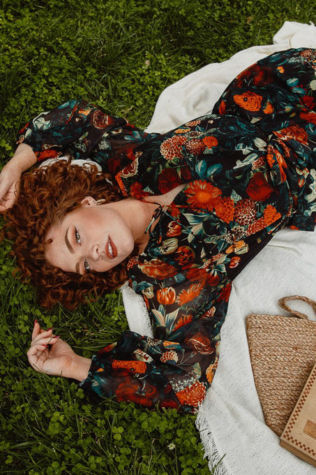A woman with curly red hair lounges on green grass, smiling softly in the maternity-friendly Imogen Dress in Moody Floral by Ivy City Co. A woven bag is placed nearby.