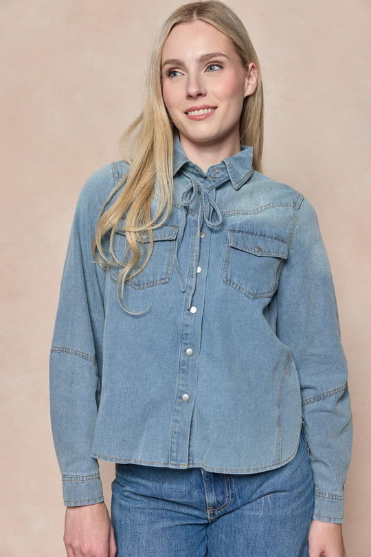 A woman with long blonde hair wears the Denver Top in Lightwash Denim by Ivy City Co, a western-inspired blouse featuring front pockets and pearl snap buttons, paired with jeans. She stands against a beige background, smiling slightly off camera.