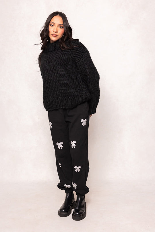 A woman with long dark hair wears a chunky black knit sweater, Ivy City Co’s Micah Jogger Sweatpants in Black featuring white bow designs, and black boots, standing against a light background.