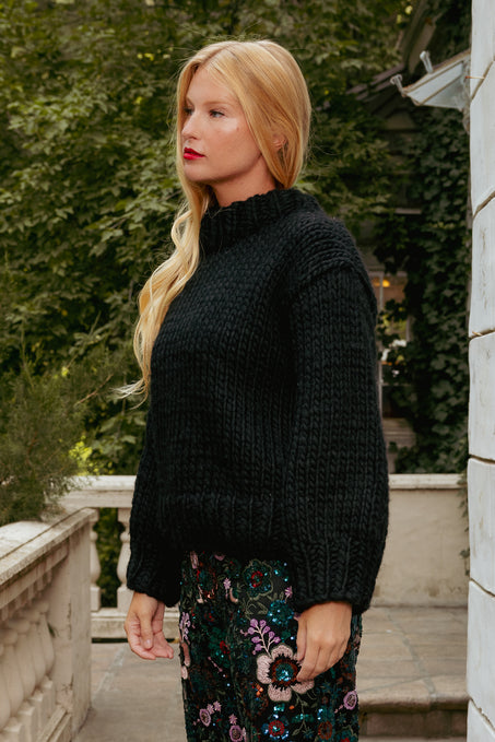Woman wearing a black sweater and floral pants standing outdoors.