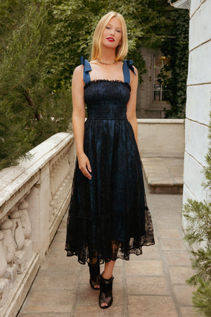 Woman in a black dress standing outdoors with greenery and stone steps in the background