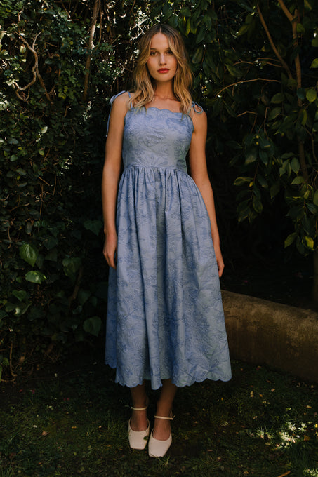 A woman with wavy blond hair stands outside before green foliage, wearing the Ivy City Co Isla Dress in Floral Embroidered Chambray and white shoes. The sleeveless dress features a fitted bodice and full skirt.