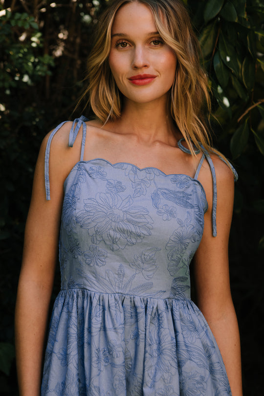A woman with wavy blonde hair stands outdoors before leafy greenery, smiling softly at the camera in the Ivy City Co Isla Dress in Floral Embroidered Chambray with tie straps and a maternity-friendly fit.