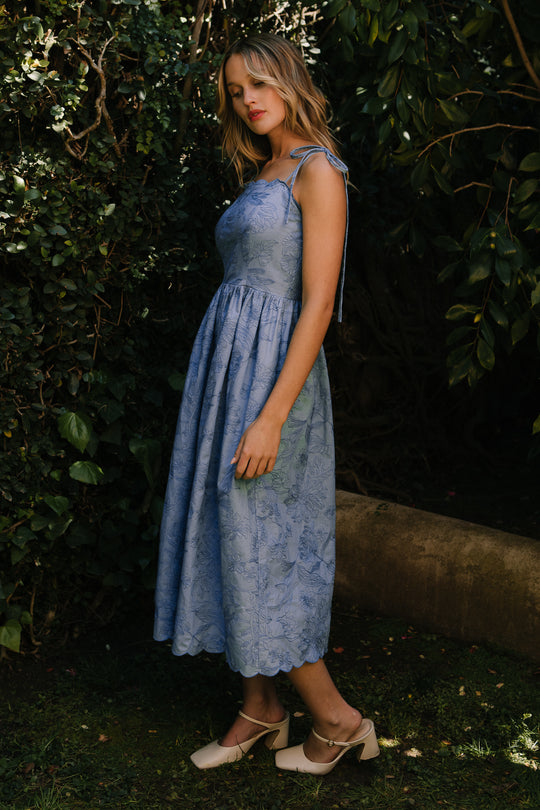 A woman stands outdoors in the Ivy City Co Isla Dress in Floral Embroidered Chambray, a blue sleeveless midi dress with tie straps and beige heels, surrounded by green foliage and trees.
