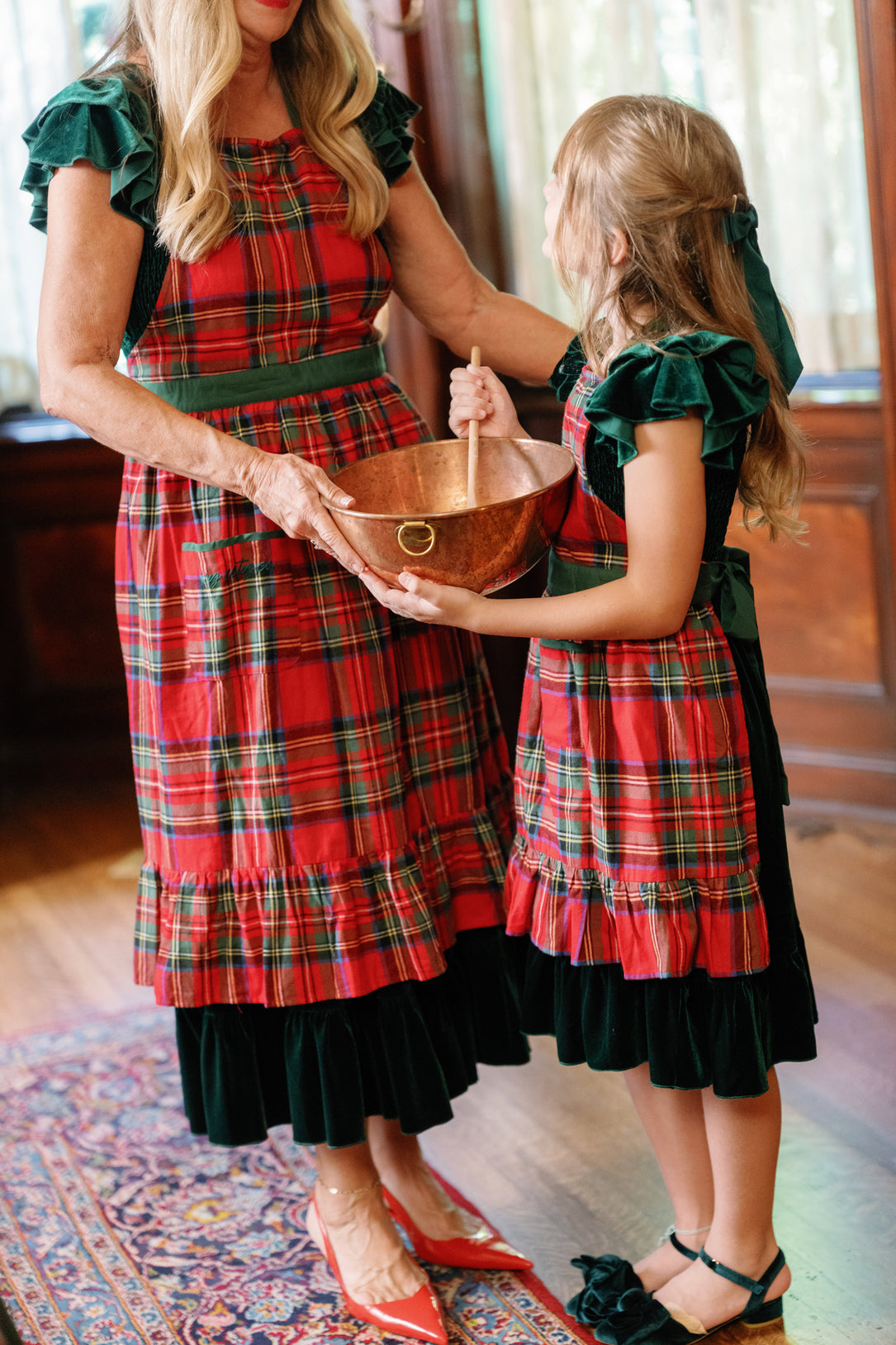 An adult and a child, both wearing Ivy City Co’s Ivy Apron in Holiday Plaid, stand together in a warmly lit room, holding and stirring a copper mixing bowl. Their cozy matching aprons add a festive touch to their holiday baking tradition.