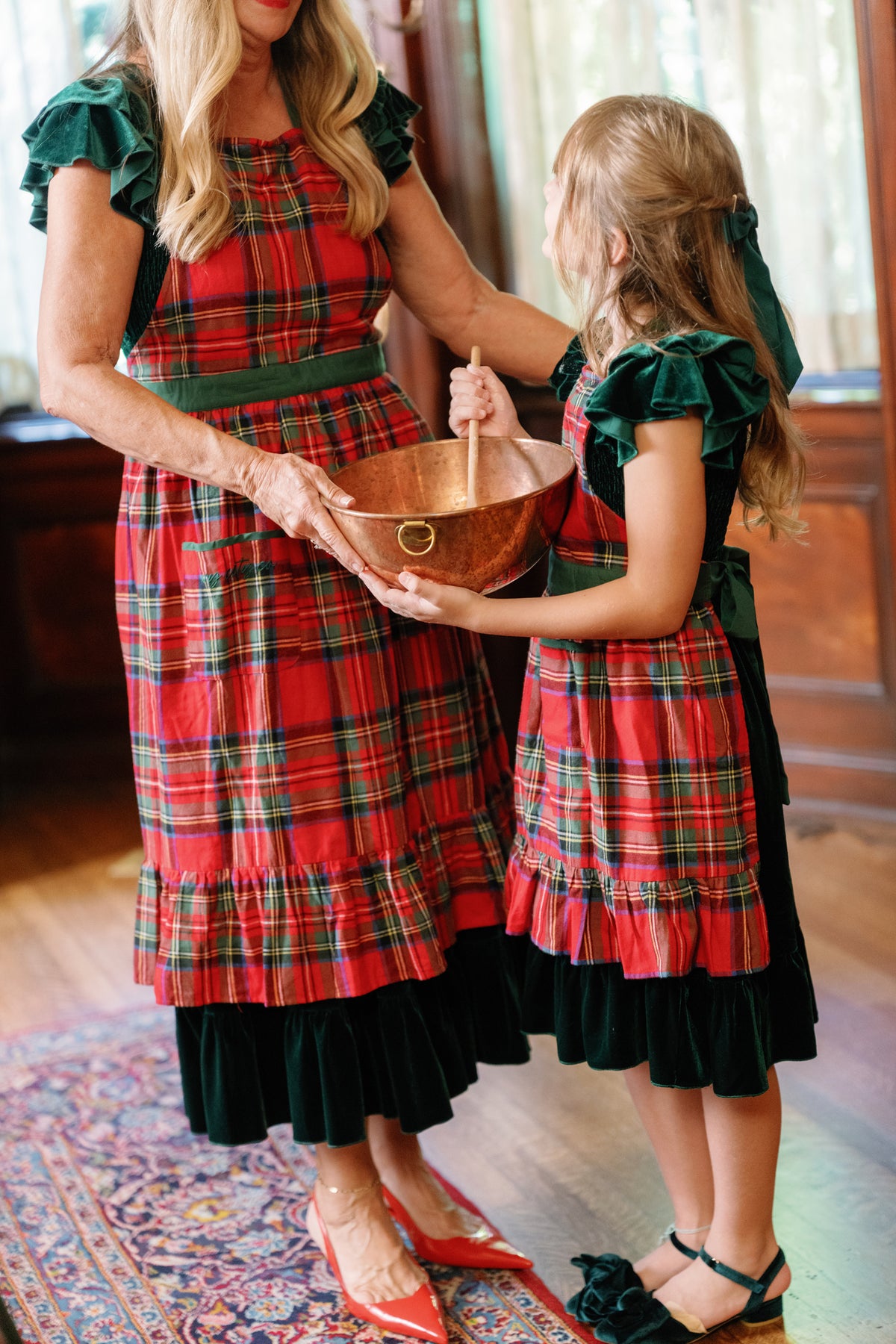 An adult and a child, both wearing Ivy City Co’s Ivy Apron in Holiday Plaid, stand together in a warmly lit room, holding and stirring a copper mixing bowl. Their cozy matching aprons add a festive touch to their holiday baking tradition.