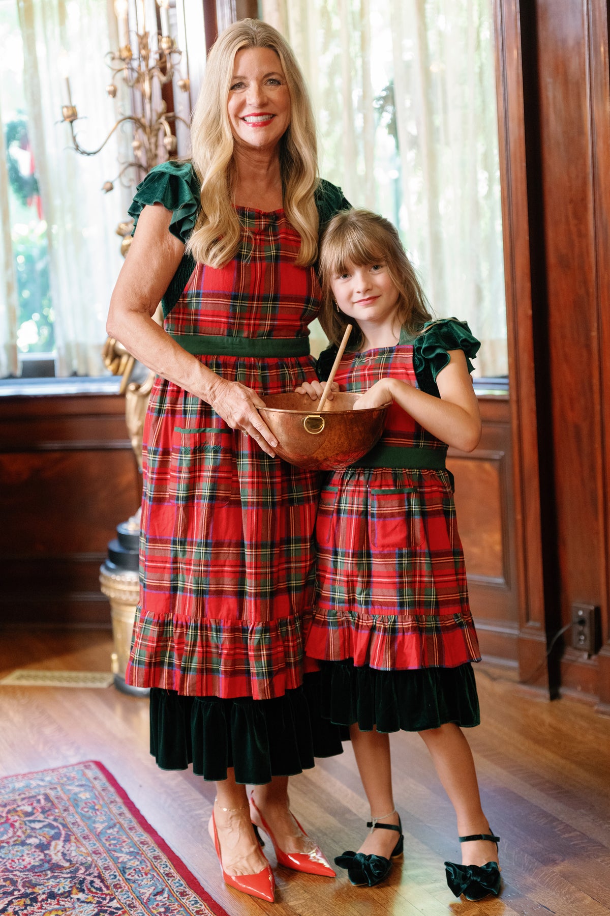 A woman and a girl, both wearing the Ivy Apron in Holiday Plaid by Ivy City Co over their dresses, stand together indoors. The woman holds a wooden mixing bowl and spoon as they smile, standing on a patterned rug with wood paneling and sheer curtains in the background.
