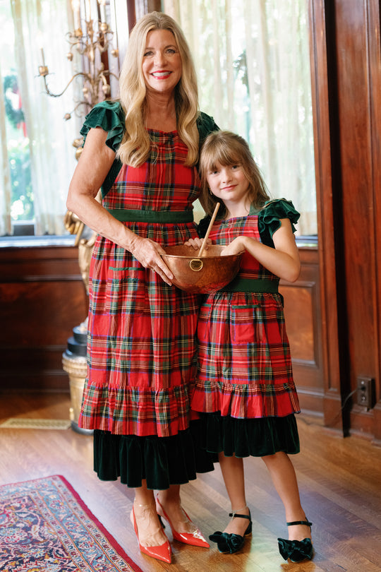 A woman and a girl, both wearing the Ivy Apron in Holiday Plaid by Ivy City Co over their dresses, stand together indoors. The woman holds a wooden mixing bowl and spoon as they smile, standing on a patterned rug with wood paneling and sheer curtains in the background.