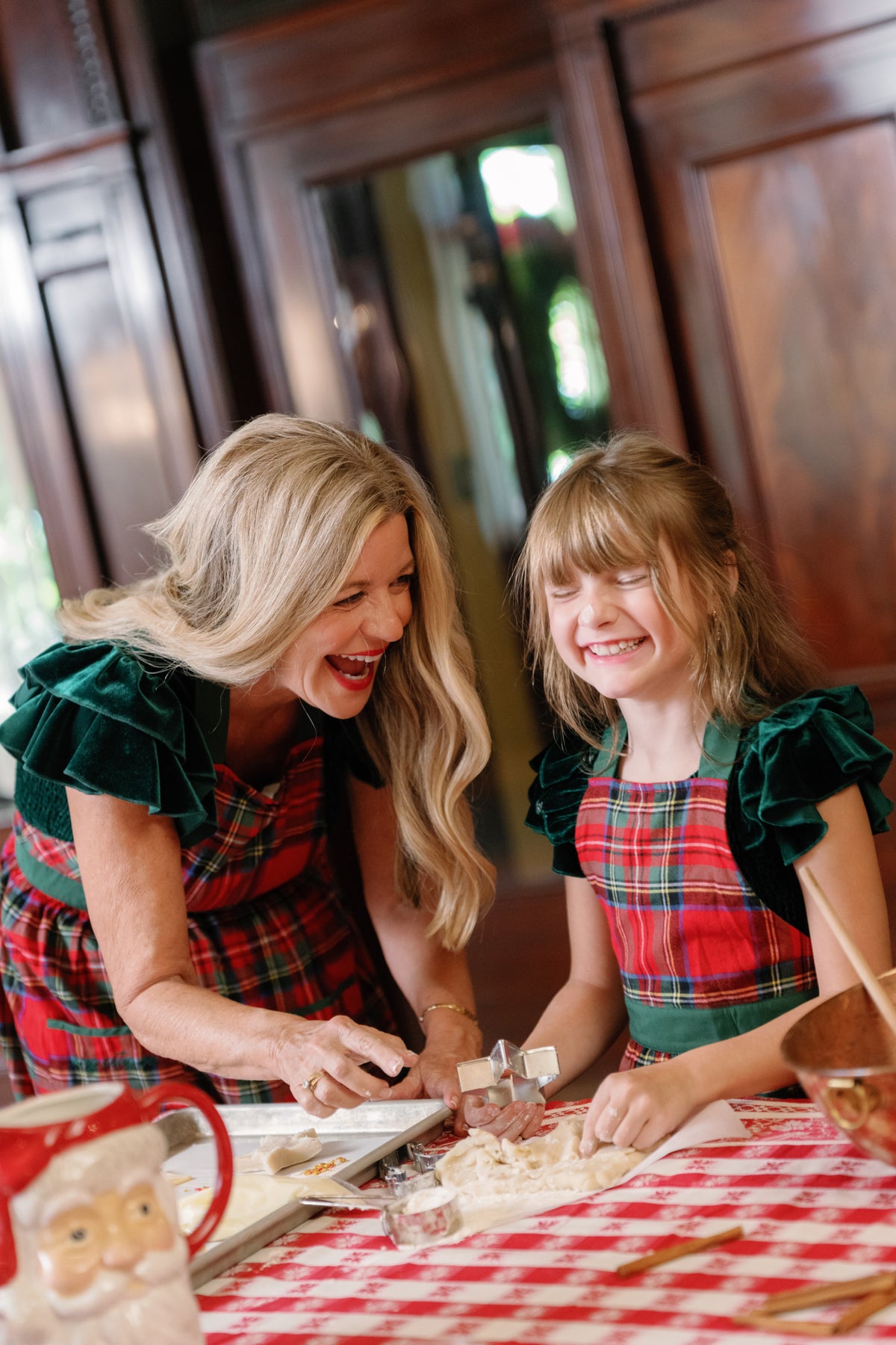 A woman and a girl, both wearing the Ivy City Co Ivy Apron in Holiday Plaid, smile as they cut cookie dough with festive cookie cutters at a table adorned with a red checked tablecloth and Christmas decorations.