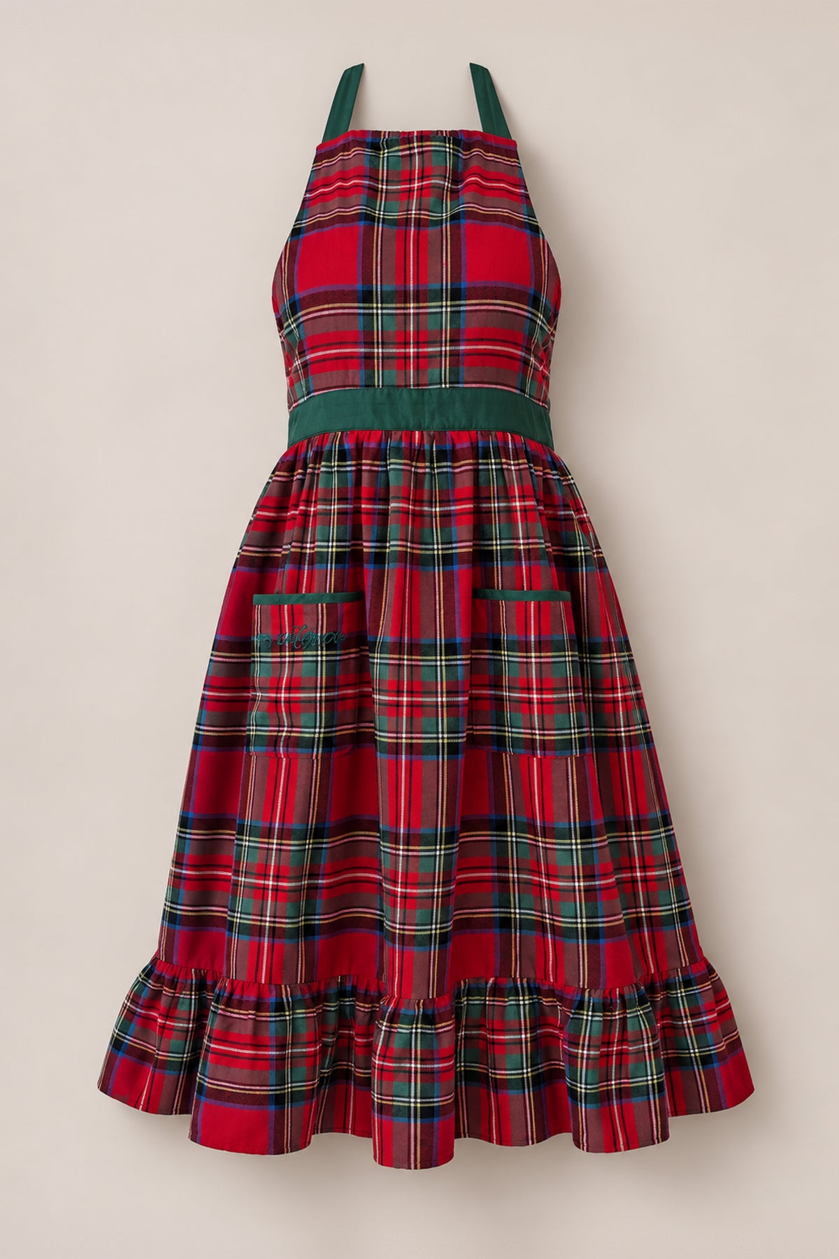 Celebrate the season in style with the Ivy Apron in Holiday Plaid from Ivy City Co, showcasing a festive red plaid pattern accented by green and yellow stripes. Thoughtfully designed with two front pockets, a green waistband, halter neck straps, and a ruffled hem, this Christmas dinner apron is set against a plain beige background for a classic holiday touch.