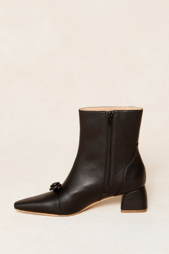 The Ivy Bow Ankle Boot by Ivy City Co is a black leather ankle boot with a side zipper, square toe, front decorative buckle, and chunky low heel, shown against a plain light background for an edgy look.