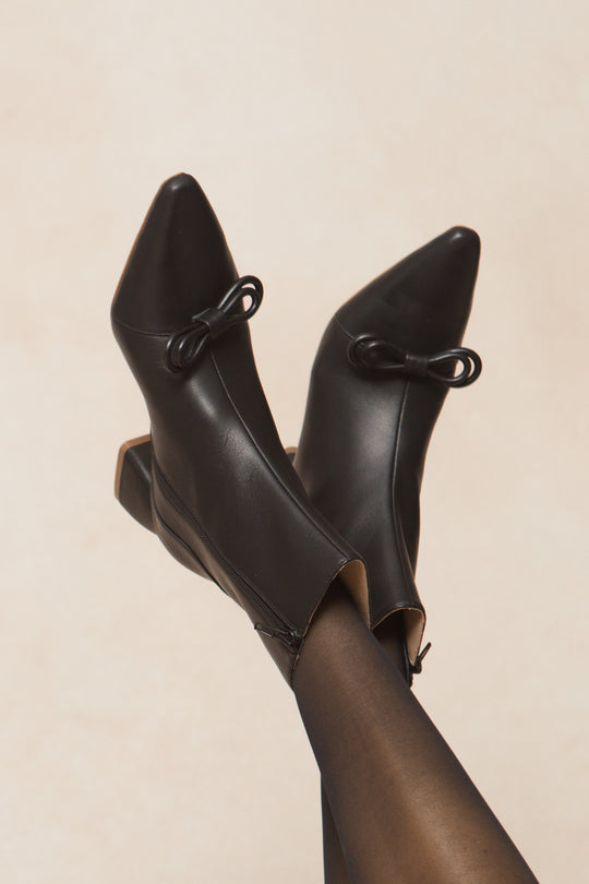 Wearing sheer black tights and Ivy City Cos Ivy Bow Ankle Boot, featuring elegant bow detailing, the person sits with legs crossed against a neutral background.