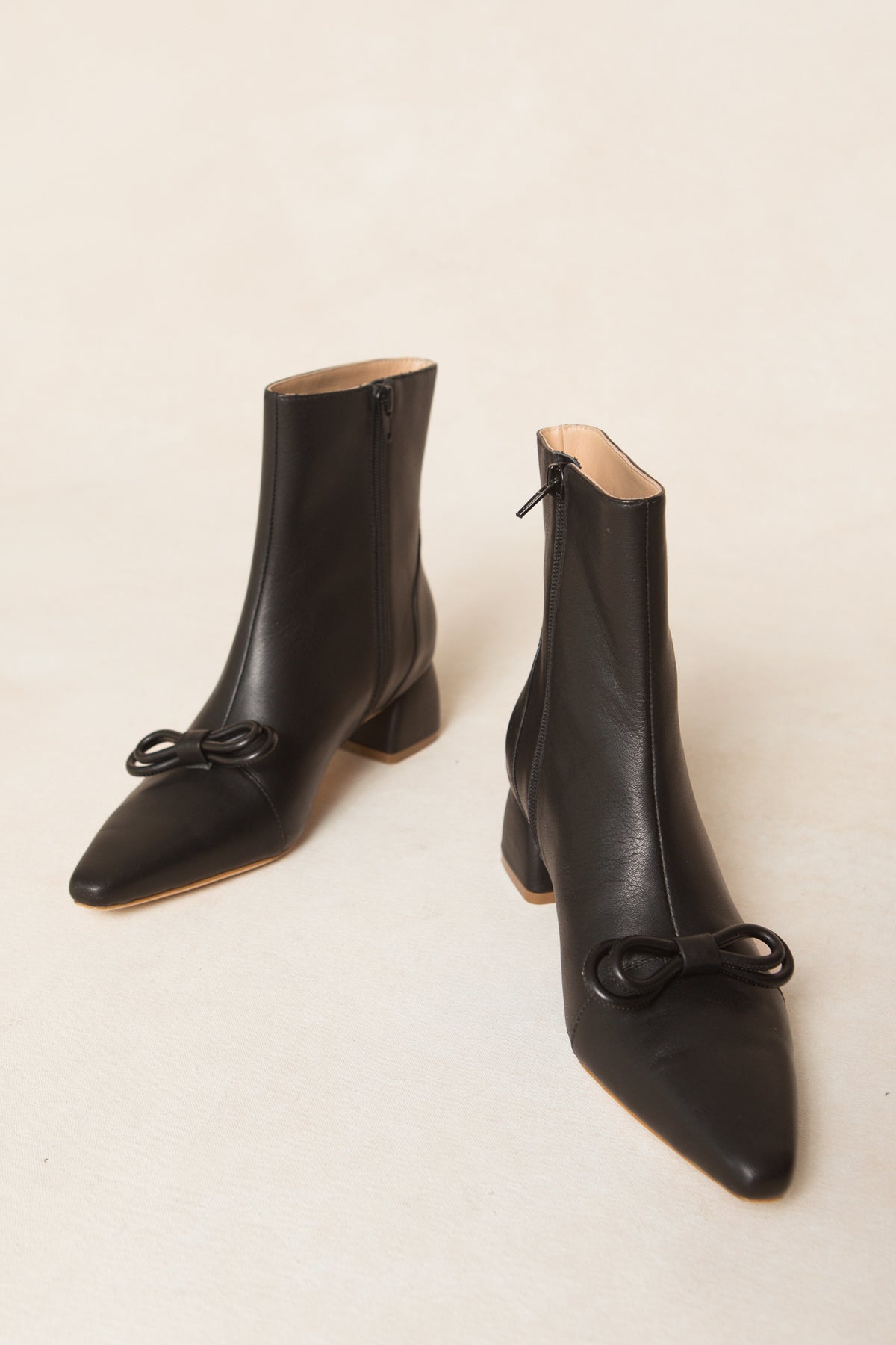 The Ivy Bow Ankle Boot by Ivy City Co features black leather, mid-heels, side zippers, and elegant bow details on pointed toes, shown against a neutral background.