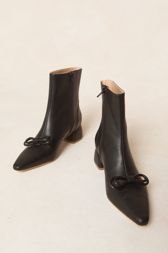 The Ivy Bow Ankle Boot by Ivy City Co features black leather, mid-heels, side zippers, and elegant bow details on pointed toes, shown against a neutral background.
