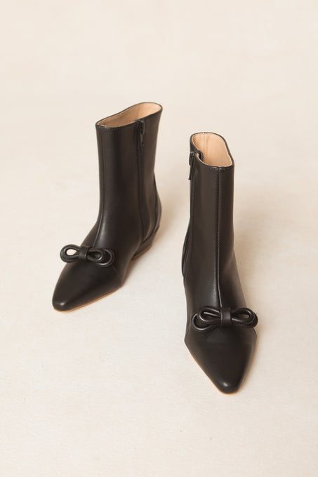 The Mini Ivy Bow Ankle Boot by Ivy City Co is a pair of black leather, ankle-high boots with a pointed toe and delicate bow detailing on the front, displayed against a light beige background.
