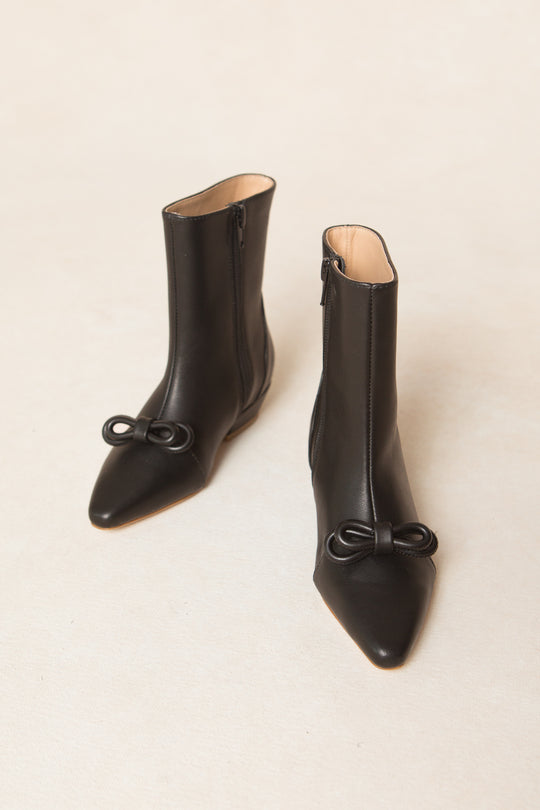 The Mini Ivy Bow Ankle Boot by Ivy City Co is a pair of black leather, ankle-high boots with a pointed toe and delicate bow detailing on the front, displayed against a light beige background.