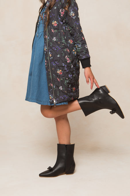 A person in a blue dress and black floral jacket balances on one leg, wearing Ivy City Co’s Mini Ivy Bow Ankle Boots in black leather with a low heel. The photo is cropped above the shoulders.