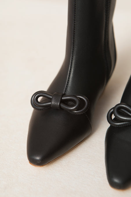 Close-up of the Mini Ivy Bow Ankle Boot by Ivy City Co, featuring black leather, a pointed toe, and an elegant bow detail on the front, set against a light beige background.
