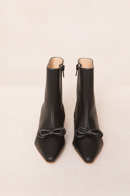 The Mini Ivy Bow Ankle Boot by Ivy City Co features black leather, pointed toes, front zippers, and chic bow accents at the top, showcased against a light beige background.