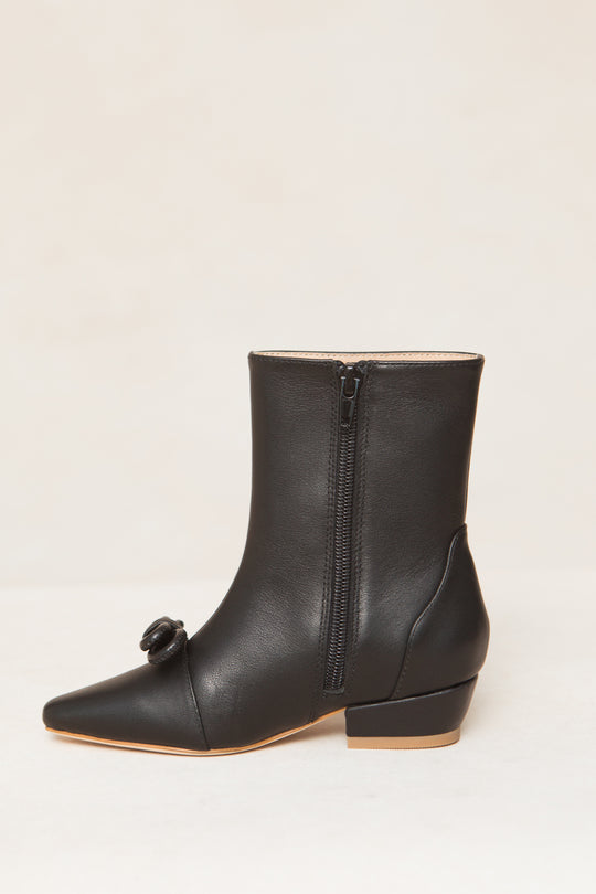 The Mini Ivy Bow Ankle Boot by Ivy City Co is a black leather boot with a low heel, pointed toe, side zipper, and a decorative bow near the toe, shown on a neutral background.