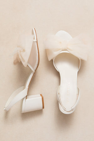 A pair of white Ivy City Co Ivy Bow Heels featuring ankle straps and large sheer tulle bows on the front, displayed on a light beige surface—perfect statement shoes for standout occasions.