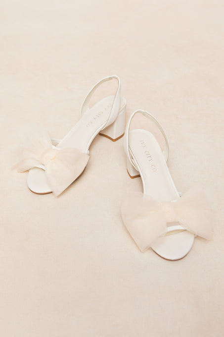 A pair of Ivy Bow Heels by Ivy City Co, featuring white open-toe design, low heels, and large sheer tulle bows on the front straps, displayed on a soft cream background.