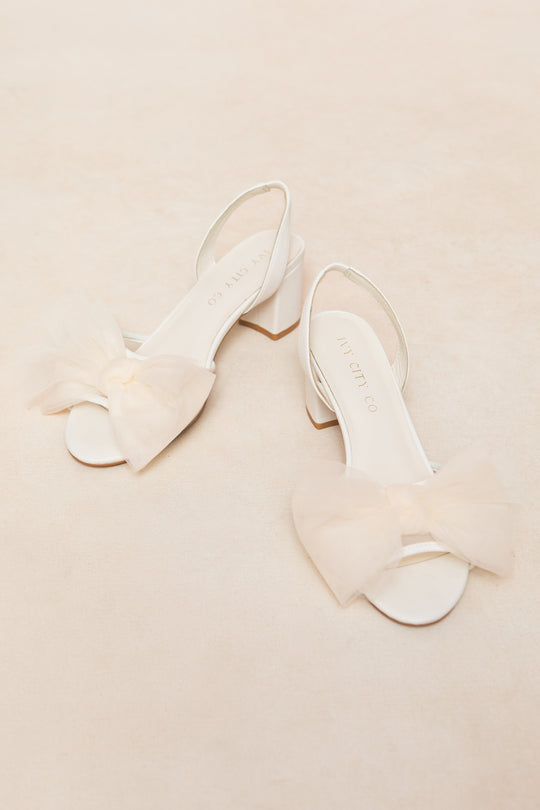A pair of Ivy Bow Heels by Ivy City Co, featuring white open-toe design, low heels, and large sheer tulle bows on the front straps, displayed on a soft cream background.