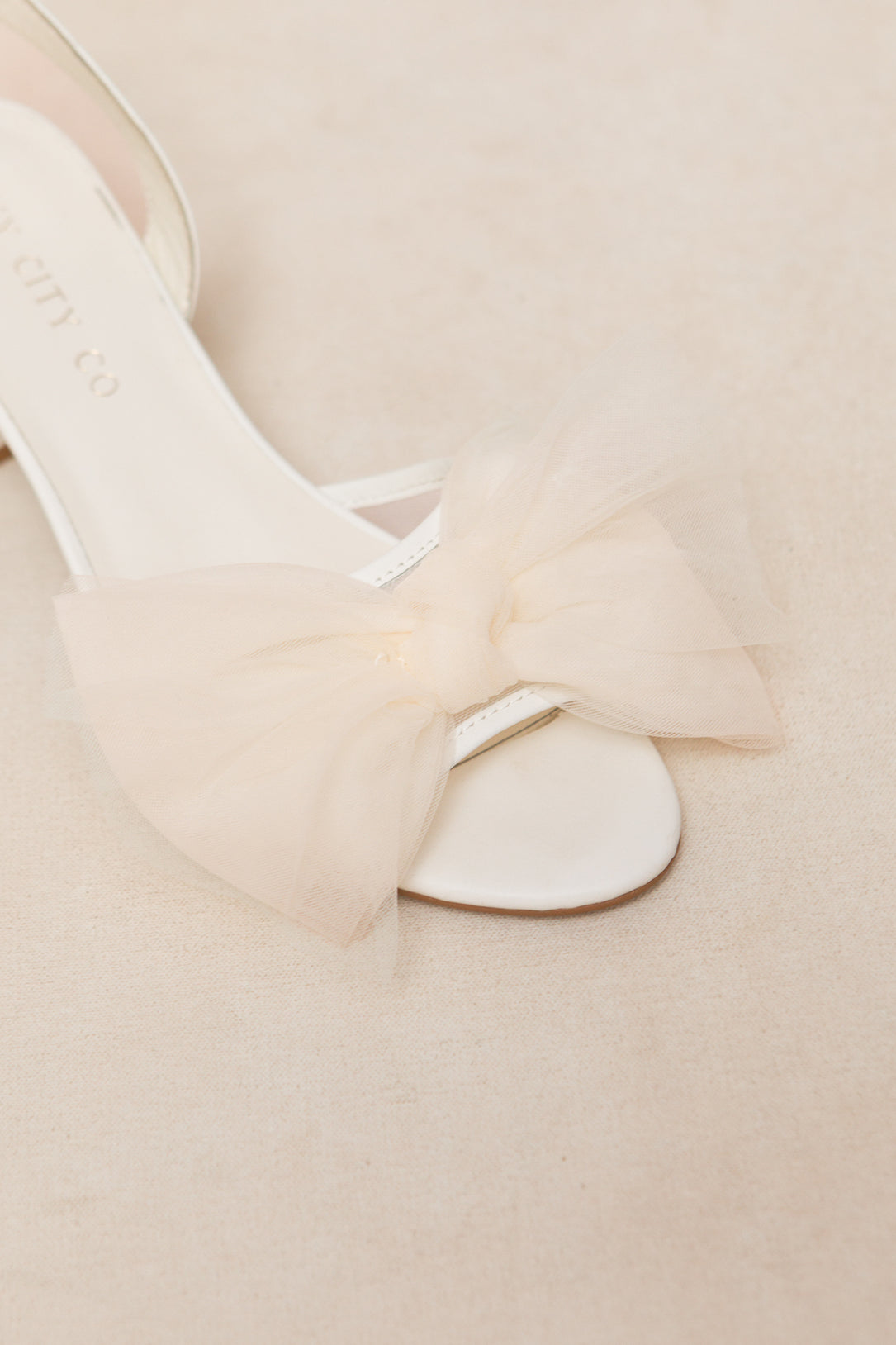 A close-up of Ivy City Cos Ivy Bow Heel, a white open-toe sandal with a large tulle bow on the strap, set against a soft beige background.