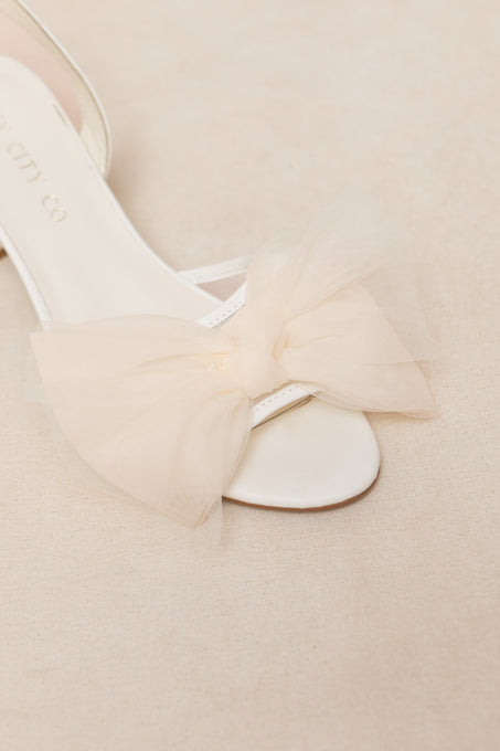 A close-up of Ivy City Cos Ivy Bow Heel, a white open-toe sandal with a large tulle bow on the strap, set against a soft beige background.
