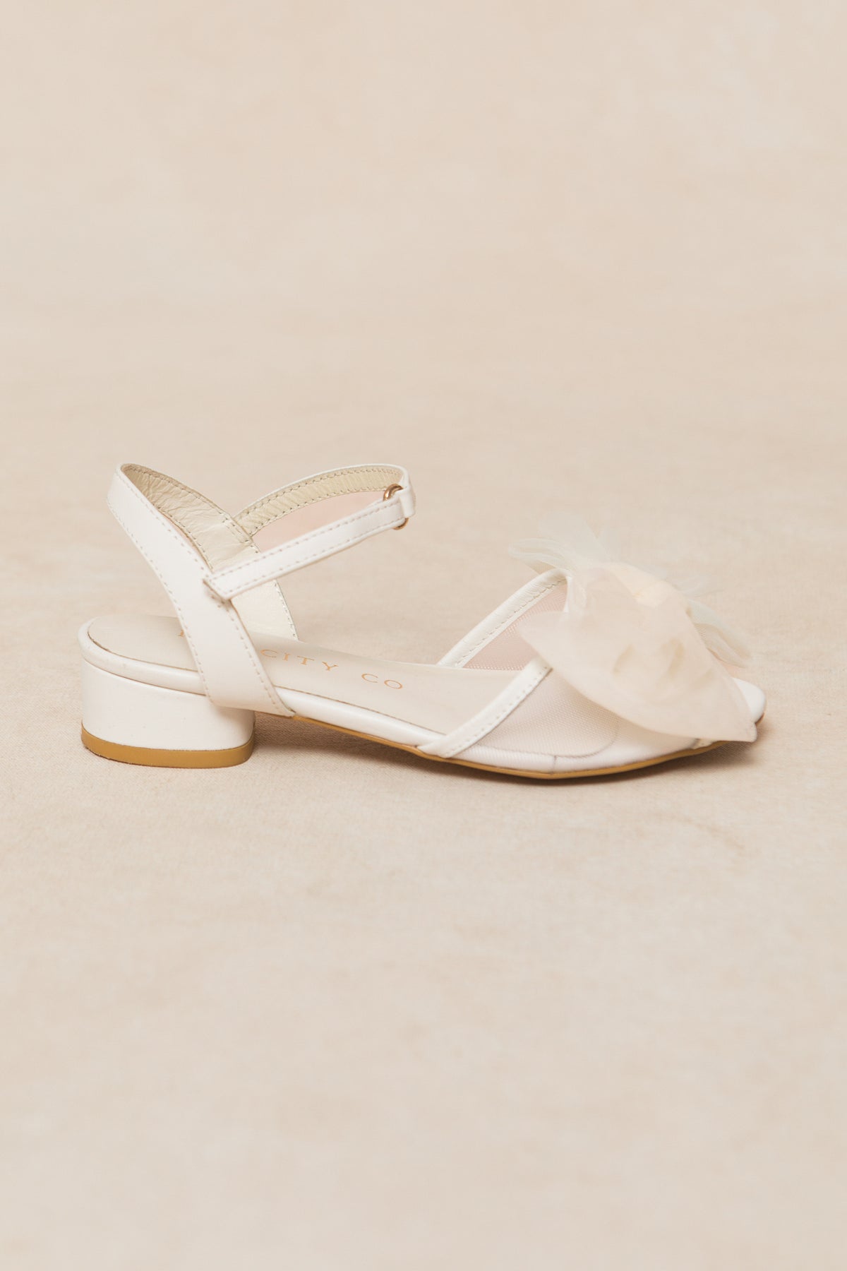 The Ivy City Co Mini Ivy Bow Heel in White, featuring a low block heel, ankle strap, and a large sheer tulle bow on the front, is displayed against a light beige background.