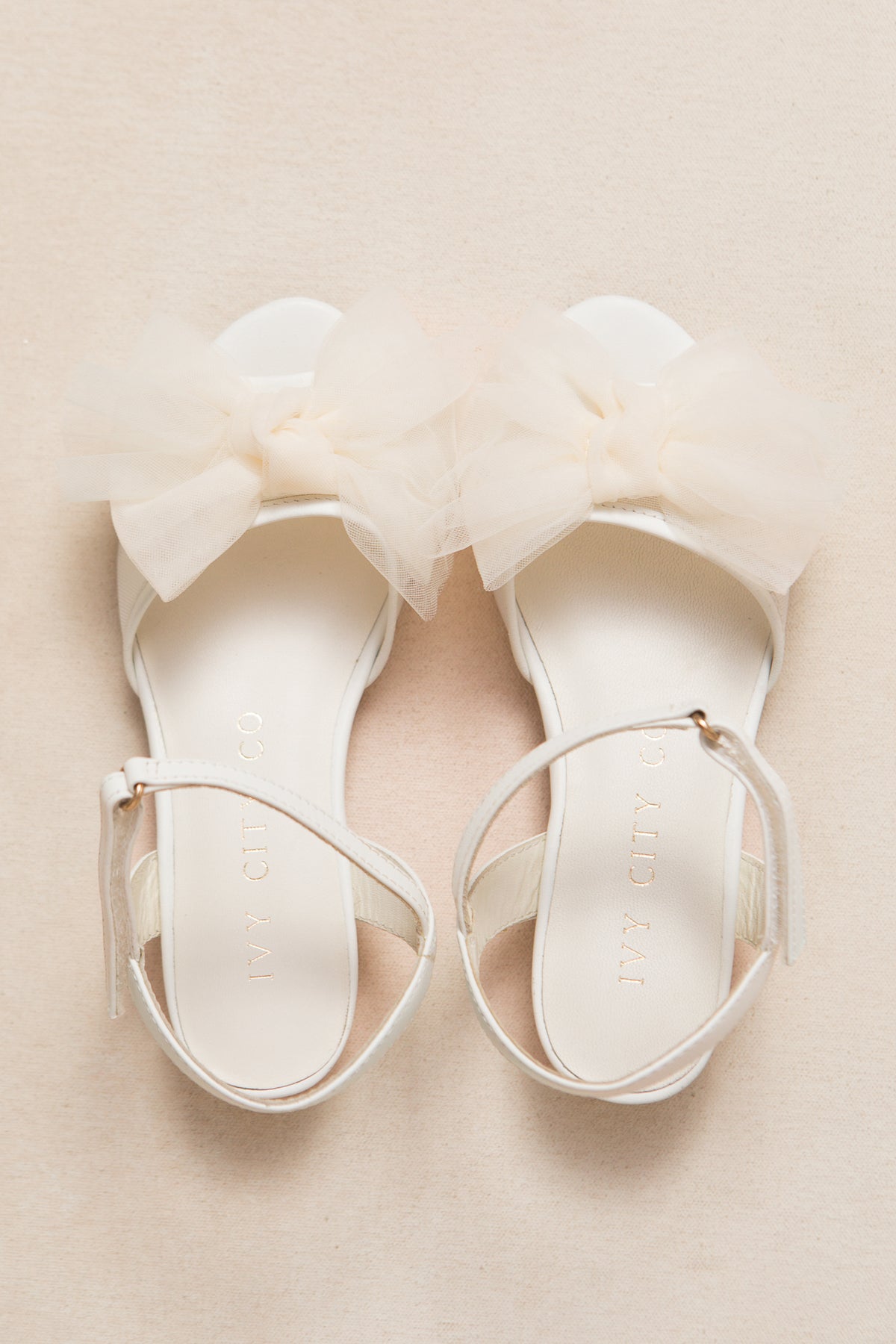The Mini Ivy Bow Heel in White by Ivy City Co features elegant ankle straps and a large sheer tulle bow on the front, displayed on a light beige surface.