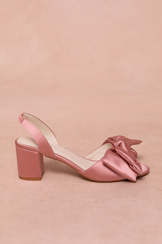 The Ivy Bow Heel in Pink Satin by Ivy City Co is a single open-toe sandal featuring a low block heel, slingback strap, and large front bow, shown against a matching pink background.