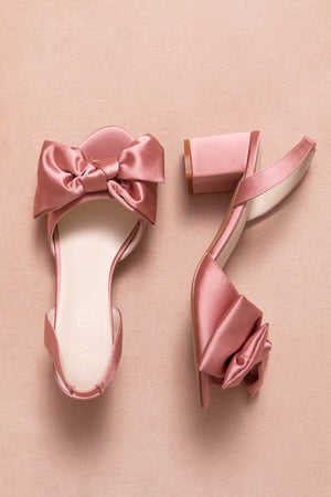 Two Ivy City Co Ivy Bow Heels in Pink Satin, featuring block heels and large decorative bows, are displayed on a soft pink background.
