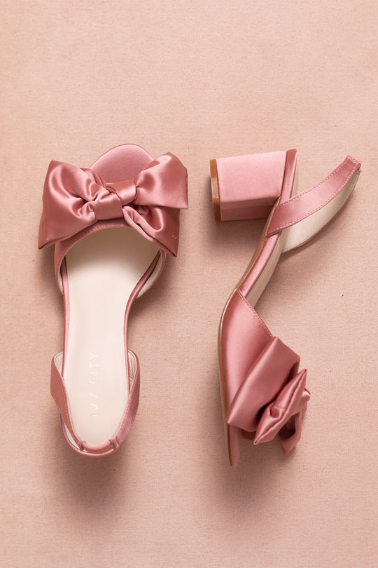 Two Ivy City Co Ivy Bow Heels in Pink Satin, featuring block heels and large decorative bows, are displayed on a soft pink background.