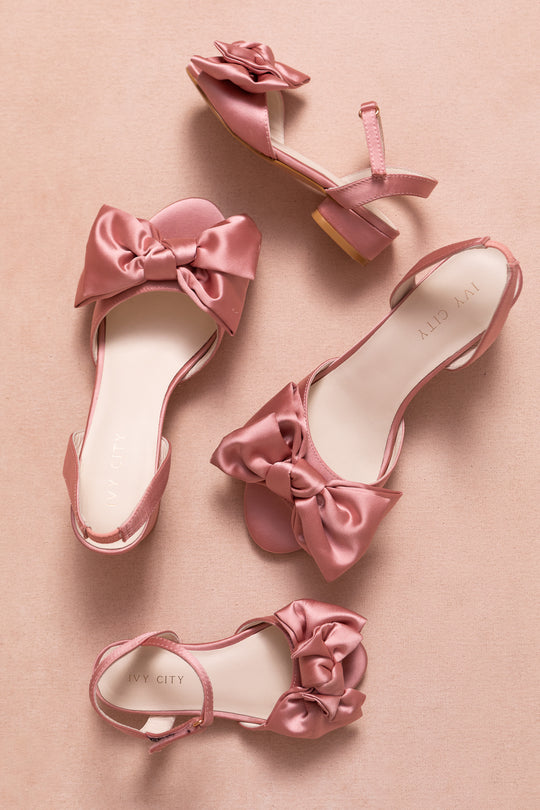 Four Ivy Bow Heels in Pink Satin by Ivy City Co, featuring large bows, ankle straps, low heels, and IVY CITY footbeds, are arranged on a soft pink background for an elegant touch.