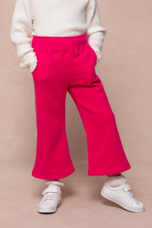 A child wearing Ivy City Cos Mini Ivy Jewel Heart Sweatpants in Pink, a white ribbed sweater, lace-trimmed socks, and white sneakers stands against a beige background—perfect for girls seeking comfy style.