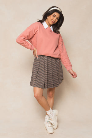 A woman poses against a beige background wearing the Ivy City Co Ivy Sweatshirt in Pink, styled oversized over a white collared shirt, with a patterned pleated skirt, white socks, sneakers, and a white headband.