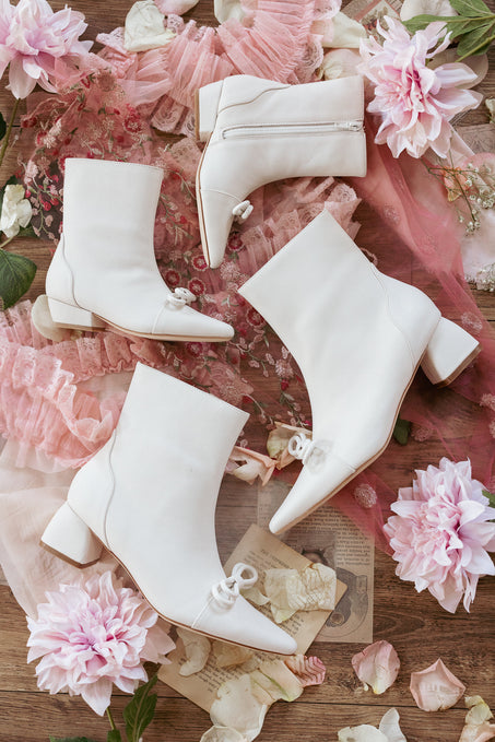 Four pairs of Ivy City Co’s Mini Ivy Bow Ankle Boot in White are arranged on a wooden surface, surrounded by pink lace, vintage papers, and pink flowers.