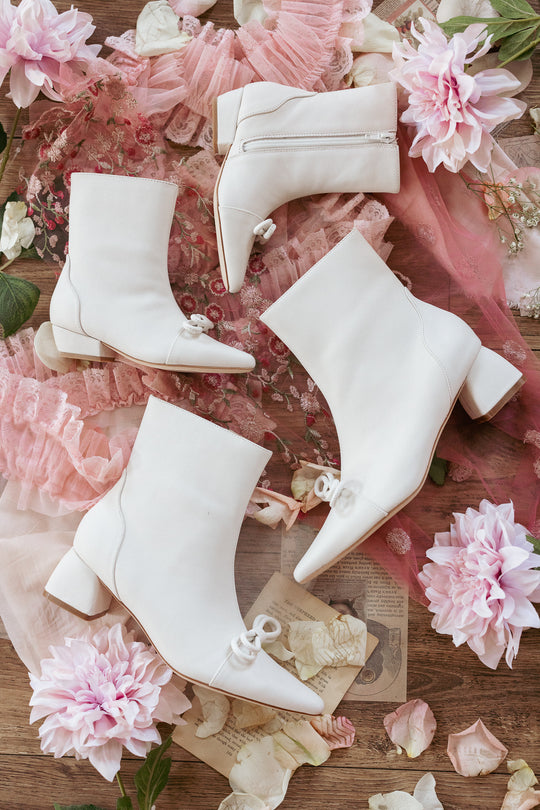 Four pairs of Ivy City Co’s Mini Ivy Bow Ankle Boot in White are arranged on a wooden surface, surrounded by pink lace, vintage papers, and pink flowers.