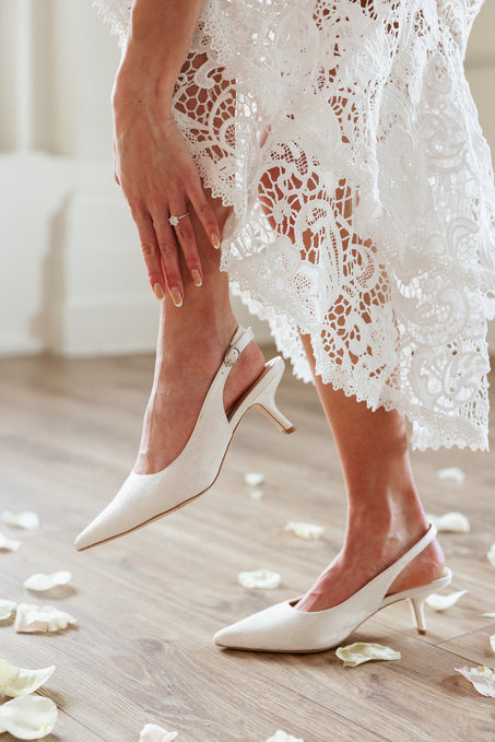 A woman in a white lace dress adjusts her Ivy City Co Ivy Lace Kitten Heel in White shoes, standing on a wooden floor covered with white rose petals. Her engagement ring gleams on her left hand.