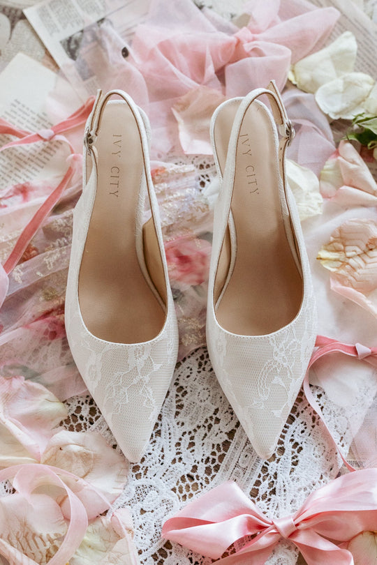 The Ivy Lace Kitten Heel in White from Ivy City Co rests on lace, surrounded by pink ribbons, floral petals, and book pages—an elegant choice for wedding shoes.