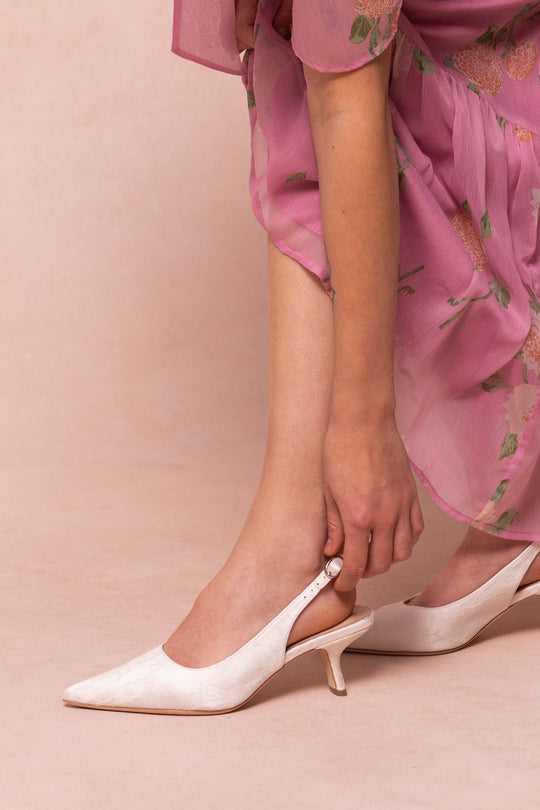 A person in a pink floral dress adjusts the strap of the Ivy Lace Kitten Heel in White by Ivy City Co. Only their lower body and hand are seen against a soft beige background, showcasing these elegant wedding shoes.