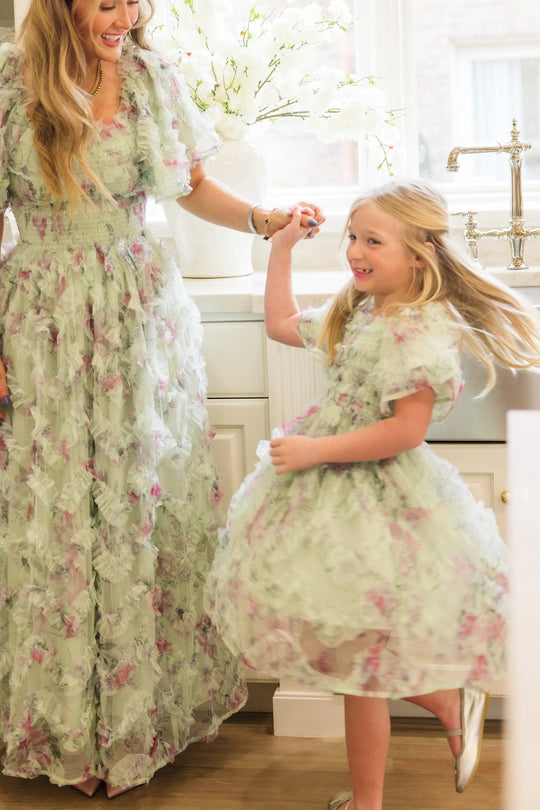 A woman and a young girl, both dressed in matching Ivy City Co Florentina Dresses in Pistachio Aquarelle Fleur, smile and hold hands as they dance together in a bright kitchen near a window adorned with white flowers. The woman’s dress is maternity friendly, offering comfort and elegance for every special moment.