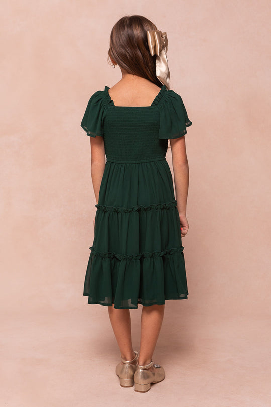 A girl seen from behind wearing an emerald chiffon Ivy City Co Mini Ellie Dress with a smocked bodice, flutter sleeves, and a tiered skirt.
